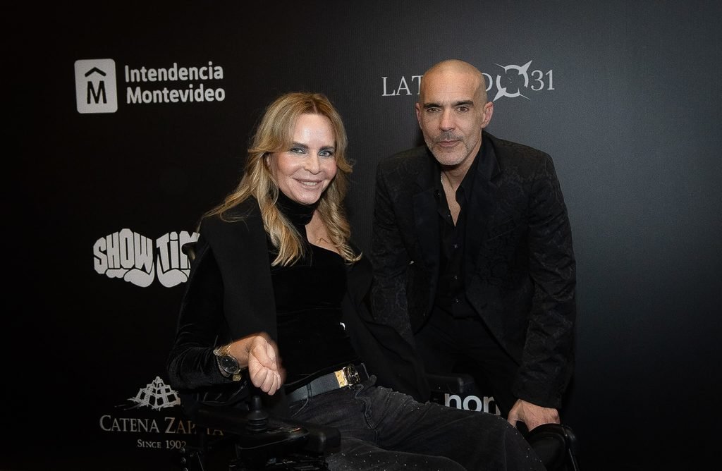 Mujer rubia y hombre de negro posan sentados frente al panel de sponsors en el Renato Papiccio Runway, Sofitel Montevideo