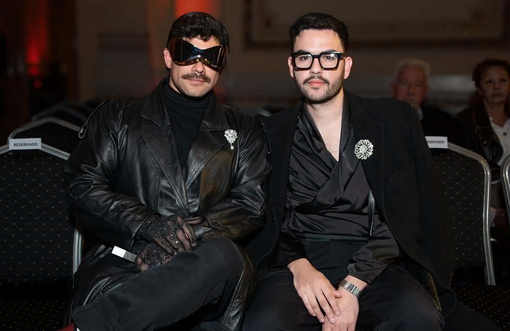 Dos invitados sentados en primera fila, uno con antifaz negro y chaqueta de cuero en el Renato Papiccio Runway, Sofitel Montevideo